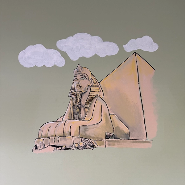 A mural depicting a sphinx seated before a pyramid, set against a pastel green background with soft, white clouds above.